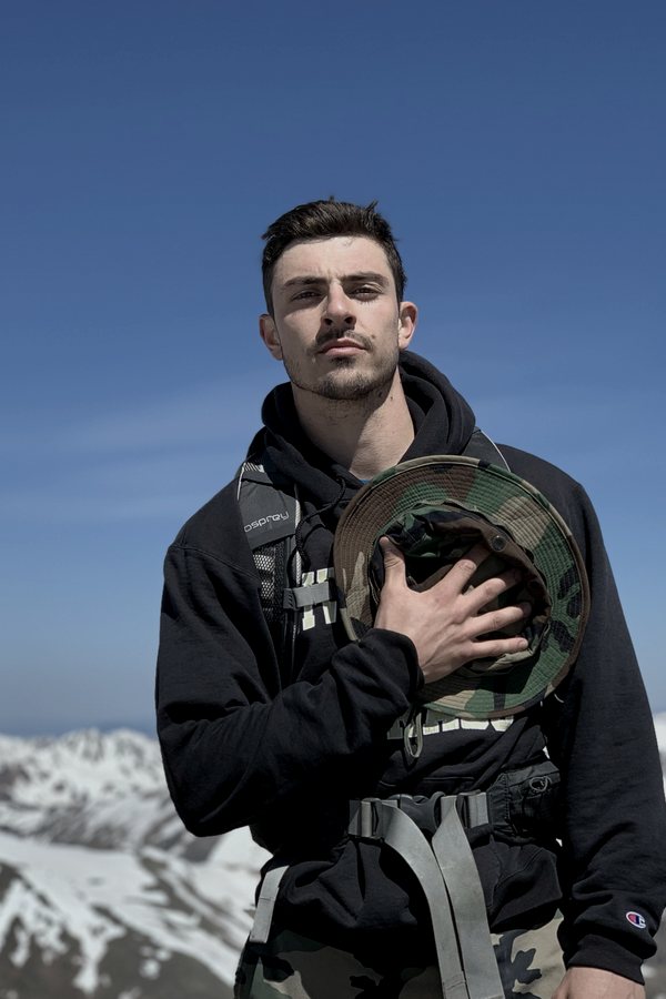 Jared on a snowy mountain summit with his pack, hand over heart holding a camo boonie hat